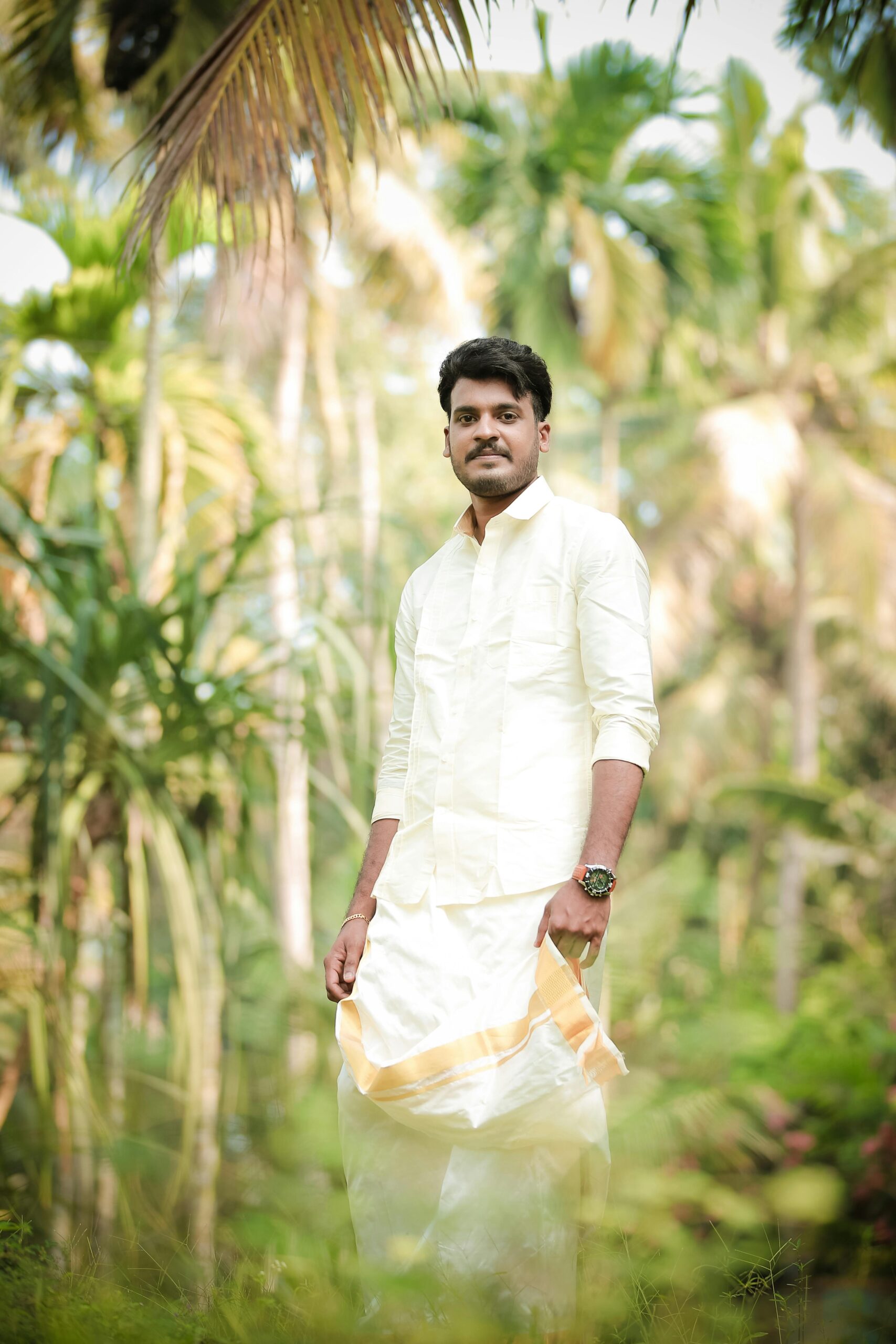 Indian groom in traditional attire posing amidst lush tropical vegetation, perfect for wedding themes.